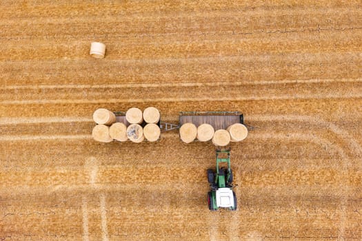 A tractor loads straw bales on two trailers in a dry field in hot summer, aerial view from Germany