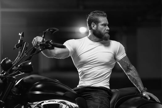 Sexy handsome attractive sporty muscular fitness model breaded biker in white tight tshirt sits on brutal beautiful motorcycle in the parking, black and white photo