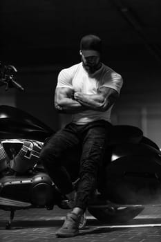 Sexy handsome attractive sporty muscular fitness model breaded biker in black cap and white tight tshirt sits on brutal beautiful motorcycle in the parking, black and white photo