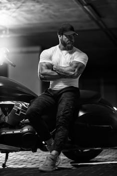 Sexy handsome attractive sporty muscular fitness model breaded biker in black cap and white tight tshirt sits on brutal beautiful motorcycle in the parking, black and white photo
