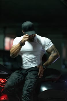Sexy handsome attractive sporty muscular fitness model breaded biker in black cap and white tight tshirt sits on brutal beautiful motorcycle in the parking