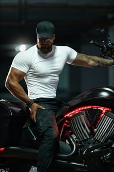 Sexy handsome attractive sporty muscular fitness model breaded biker in black cap and white tight tshirt sits on brutal beautiful motorcycle in the parking