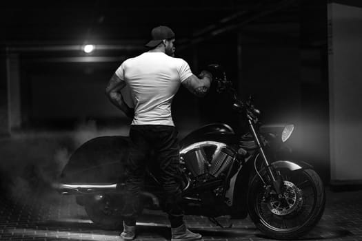 Sexy handsome attractive sporty muscular fitness model breaded biker with brutal beautiful motorcycle in the parking posing with a motorbike with his back to the frame, black and white photo