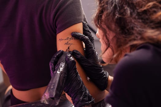 Talented focused woman tattoo artist diligently crafting personalized lettering tattoo using machine on arm of female client, cropped shot. Tattooing process concept