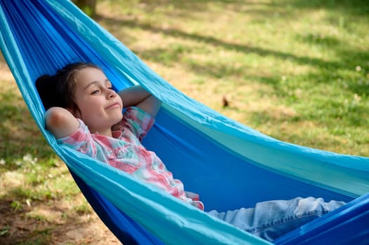 Close-up portrait of an adorable Caucasian child girl dressed in casual denim and pink checkered shirt, relaxing, lying on a blue hammock in the backyard or summer camp, enjoying her weekend outdoors