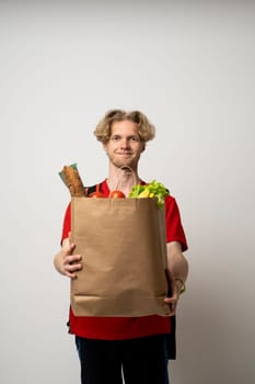 Delivery man guy employee man 20s wear red uniform work as dealer courier hold craft brown paper bag with grocery food vegetables