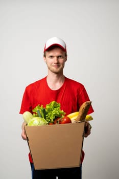 Man courier in red uniform holding paper box with fresh products, groceries, food on white background. Food delivery service