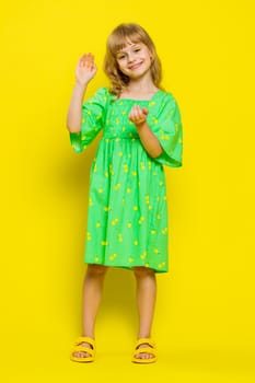 Young blonde school girl smiling friendly at camera and waving hands gesturing hello or goodbye, welcoming with hospitable expression. Preteen female child kid isolated on yellow background. Vertical