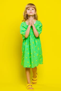 Please, God help. Blonde child girl kid praying, looking at camera making wish asking with hopeful imploring expression begging apology. Little preteen children isolated on yellow background. Vertical