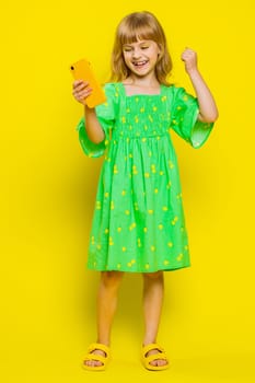Young preteen child girl kid use mobile smartphone typing browsing say Wow yes found out great big win good news celebrate doing winner gesture. Little children on studio yellow background. Vertical