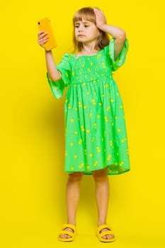 Unhappy blonde child girl kid use smartphone play online game, typing browsing, loses surprised sudden lottery results, bad fortune, loss, fail. Little preteen children on yellow background. Vertical
