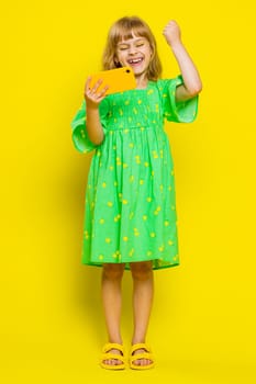 Worried funny young school girl enthusiastically playing racing or shooter video games on smartphone. Preteen child kid using smartphone gadget app with drive simulator on yellow background. Vertical