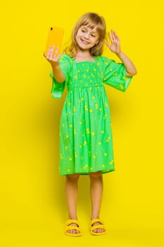 Young school girl blogger influencer taking selfie on smartphone, communicating video call online with subscribers in social media app. Preteen child kid isolated on studio yellow background. Vertical