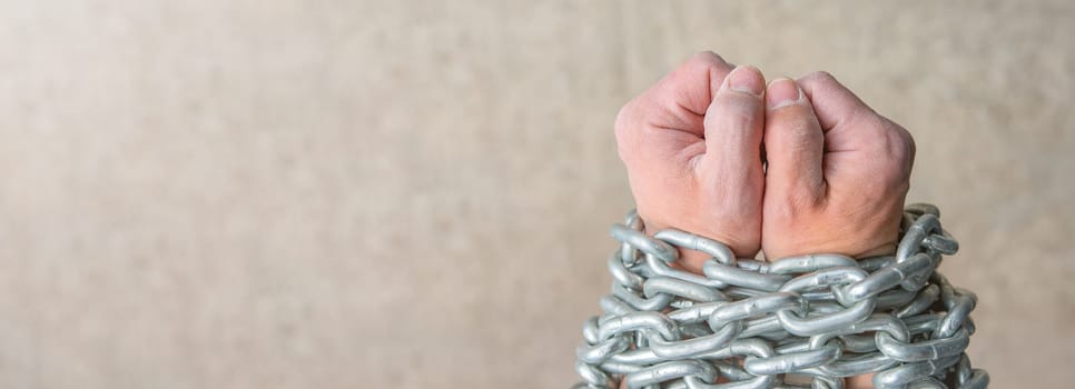 The rough hands of a worker, a white male are wrapped in a chain as a concept of slavery or imprisonment. Close-up photo of the hands of a slave with a chain on a gray background with copy space