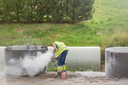 Installing a concrete pipe in the ground to collect rainwater. The worker cuts holes in the concrete part of the pipe to connect to the storm drain. A man in construction clothes is sawing concrete