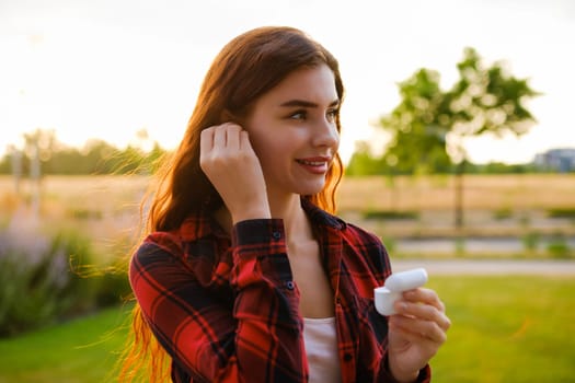 A lovely woman beams with a smile wearing plain shirt, places earphones into her ears while standing on the bustling street. Enjoy music concept.