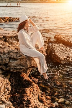 woman sea sunset. woman in a white pantsuit and hat is sitiing on the beach enjoying the sea. Happy summer holiday.