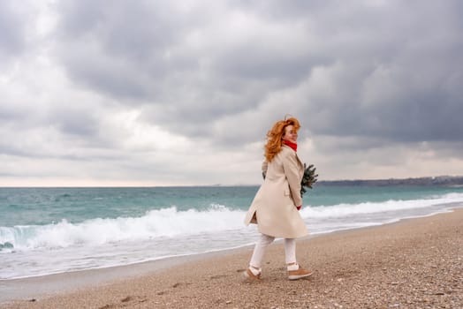 Redhead woman Christmas tree sea. Christmas portrait of a happy redhead woman walking along the beach and holding a Christmas tree in her hands. Dressed in a light coat, white suit and red mittens