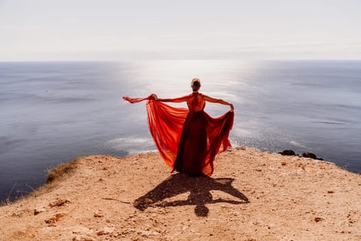 Woman red dress sea. Female dancer posing on a rocky outcrop high above the sea. Girl on the nature on blue sky background. Fashion photo