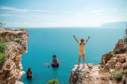 Happy girl stands on a rock high above the sea, wearing a yellow jumpsuit and sporting braided hair, depicting the idea of a summer vacation by the sea