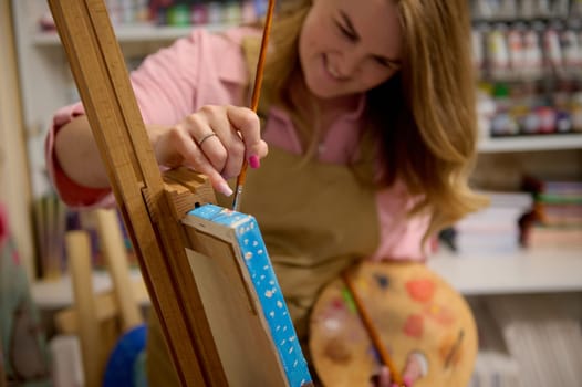 Focus on hand of blurred smiling positive female painter, inspired artist holding a paint brush, drawing picture on canvas, standing at wooden easel in her creative art studio. People. Hobby. Leisure