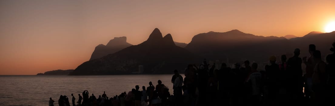 Tourists admire the sunset at Ipanema Beach, Rio de Janeiro, against a mountainous backdrop under an orange sky.