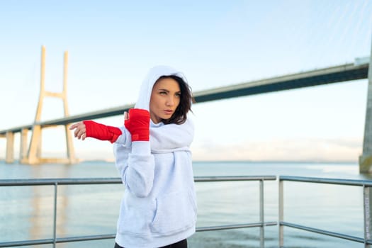 Female boxer in hoodie warming up arm muscle. Athletic female prepares for boxing workout outdoor with city seascape.Fighter warm up body before training Female fitness motivation.