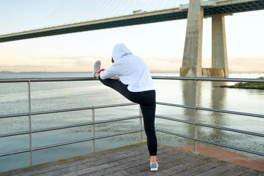 Athlete age woman stretching legs exercises. Caucasian athletic female does lunges exercises stretch leg seaside view with bridge and blue sky background. Back view