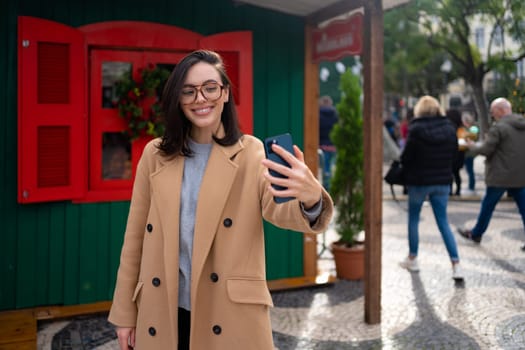 Beautiful woman using mobile phone for video call outdoors. Smiling girl looking smart phone screen in a city street. Happy smiling woman chatting on smartphone warm autumn day in European city