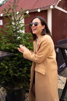 Woman looking for perfect Christmas tree to buy on the tree shop, outdoor in European city. Girl using hand for check the quality. Dressed stylish trench coat and sunglasses