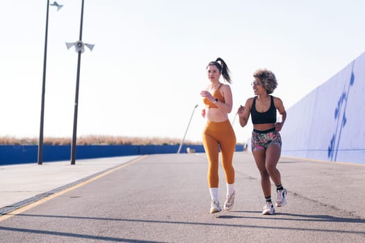 two animated female friends chatting happy while running in a athletic track in a sunny day, concept of friendship and active lifestyle, copy space for text