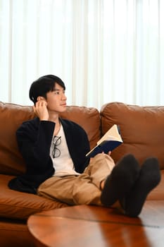 Relaxed millennial man sitting on comfortable couch and reading book, spending leisure weekend time at home..