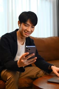 Attractive man wearing smart casual wears resting on couch and using smartphone. People and technology concept..