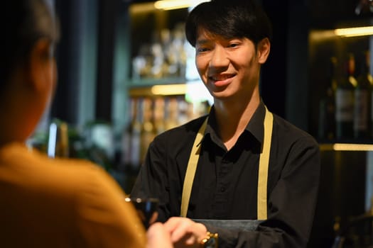Friendly man barista wearing apron standing behind counter and serving coffee to customer. Start up, small business owner, food and drink concept..