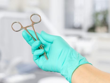 Woman's hand in a latex glove with a stainless steel needle holder. Dental office on the background.