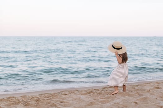Little girl in a straw hat on the beach by the sea, back view.