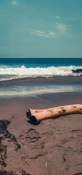 An image of Canguu Beach in Bali with a log on the sand. The log is a light brown color and has a darkened end. The sand is a light brown color and has a few small rocks scattered around. The water is a deep blue color and has white waves crashing onto the shore. The sky is a light blue color and has a few clouds scattered around. The image has a vintage filter applied to it. A log on Canguu Beach in Bali with Blue Waves- Outdoor Shot. High quality photo
