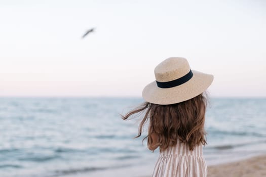 Close-up of a girl in a straw hat looks at the sea into the distance on a sunny day. The sea wind is blowing. Background of sea waves, seagulls. Pleasant little girl enjoying seascape view.