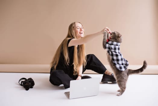 A blogger freelancer in a black is working on a laptop apple and playing, eating with Scottish straight cat. blonde assistant in video production with computer MacBook and camera Lumix GH5