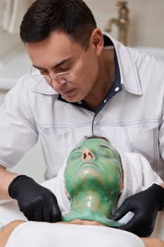 male beautician removing green alginate mask from face of beautiful woman in the spa salon. spa treatments