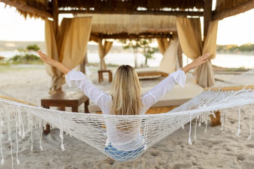The girl is resting in a hammock on the beach near the blue ocean.