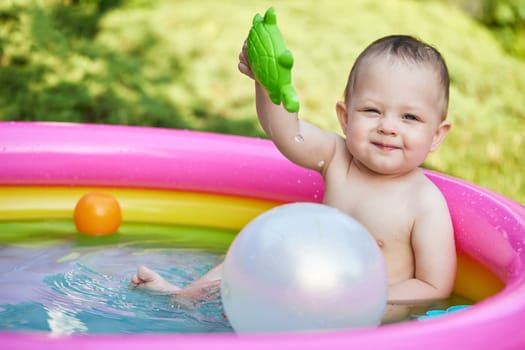 Happy cute baby girl playing with ball in kid inflatable pool in summer day in the backyard