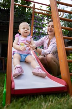 little baby girl with mother playing on slide in playground in summer. Summer family leisure.