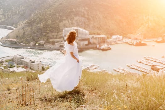 Happy woman in a white dress and hat stands on a rocky cliff above the sea, with the beautiful silhouette of hills in thick fog in the background