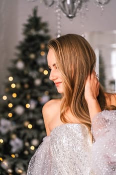 The blonde in the Christmas room. A beautiful blonde woman in a shiny light short dress with a train stands in a beautiful bright room decorated with a festive interior with a Christmas tree