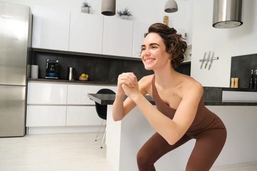 Image of young woman doing squats at home, workout on rubber mat in bright room indoors, wearing activewear for training. Copy space