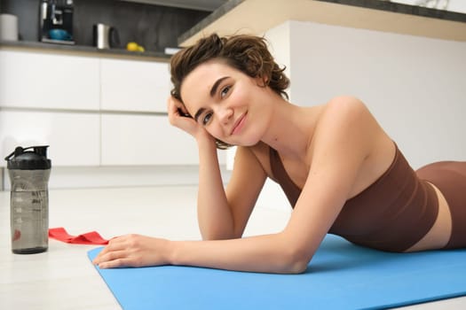 Sport and healthy lifestyle. Portrait of brunette sportswoman doing fitness in activewear, lying on rubber mat on floor, doing workout training in kitchen, drinking water from gym bottle.