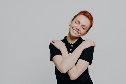 Happy red-haired woman with closed eyes dressed casually hugging herself gently and tilting head, pretty ginger female being in romantic mood while standing isolated over grey studio background