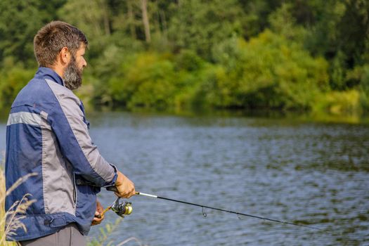 A man with a beard is fishing on the river. A fisherman with a fishing rod is fishing on the river bank.