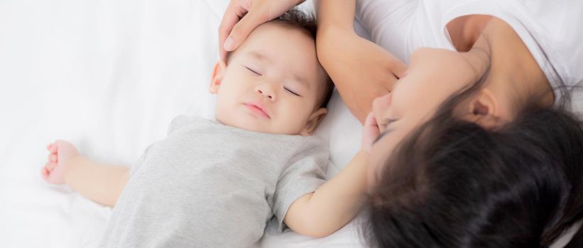 Young asian mother touch head of little baby girl with tender on bed in the bedroom, mom love newborn and care, woman with expression with child together, parent and daughter, family concept.